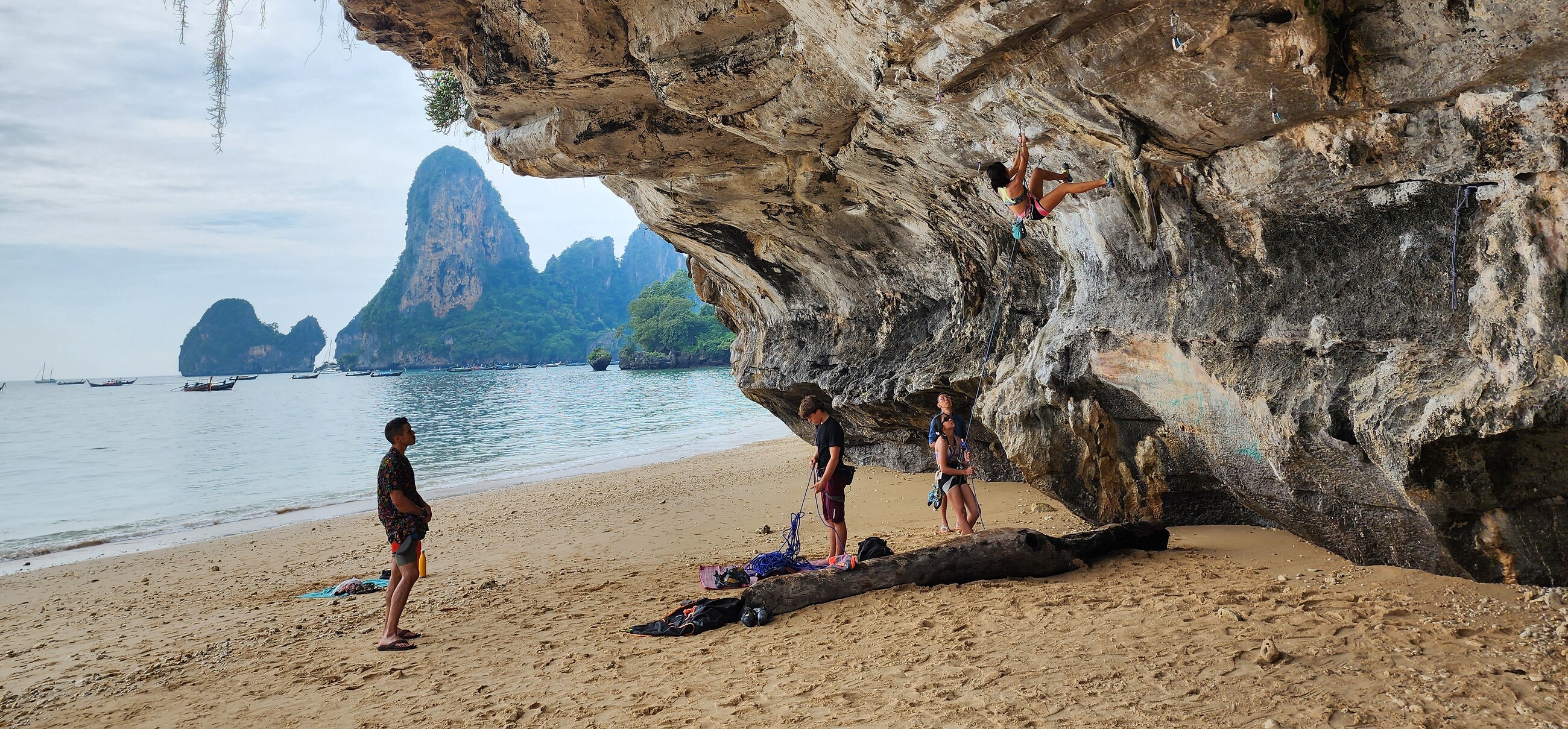 A climber on an overhanging limestone cave route in Railay, Thailand, with the belayer and others watching from the beach below and karst islands in the background
